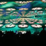 Crowd enjoying a vibrant, kaleidoscopic light show with swirling patterns above.