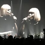Two singers with blonde bob haircuts performing on stage with microphones.