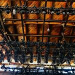 Rows of black stage lights mounted on ceiling beams in a theater.