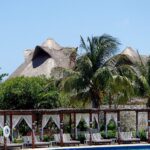 Thatched-roof huts behind tropical trees near a resort pool.