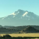 Snow-capped mountain towering over green fields and hills under a clear sky.