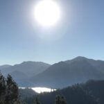 Bright sun shining over mountain range with a reflective lake below.