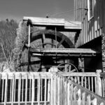 Black and white photo of an old wooden waterwheel beside a building.