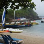 Boats docked near a sandy beach with lounge chairs and trees.