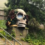 A skull sculpture adorned with flowers and sunglasses in a garden.
