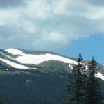 Snow patches on a mountain with trees in the foreground under a cloudy sky.