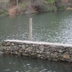 A stone wall partially submerged in a calm lake with a wooden post.